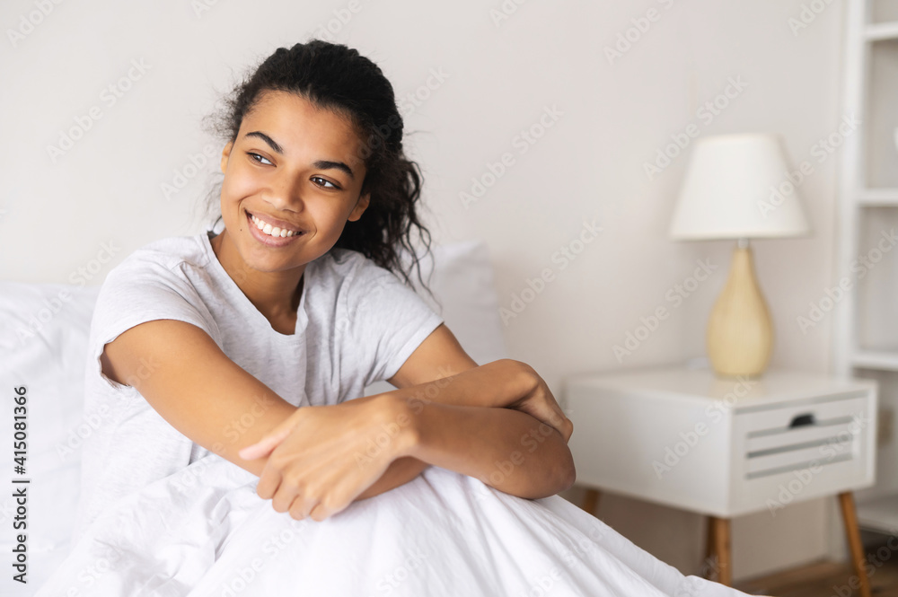 Positive young relaxed mixed-race woman covered with cozy warm soft blanket sitting down in her pajama and staying at home, daydreaming, resting and looking away, enjoying day off or a weekend in bed Positive young relaxed mixed-race woman covered with cozy warm soft blanket sitting down in her pajama and staying at home, daydreaming, resting and looking away, enjoying day off or a weekend in bed