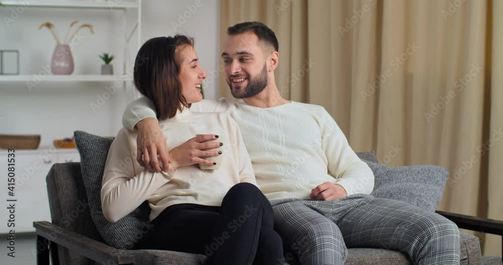 Caucasian couple young family of young women sitting at home on modern sofa in cozy living room, woman drinking tea coffee man dreaming planning shows hand gesture in air presenting imagining future Caucasian couple young family of young women sitting at home on modern sofa in cozy living room, woman drinking tea coffee man dreaming planning shows hand gesture in air presenting imagining future