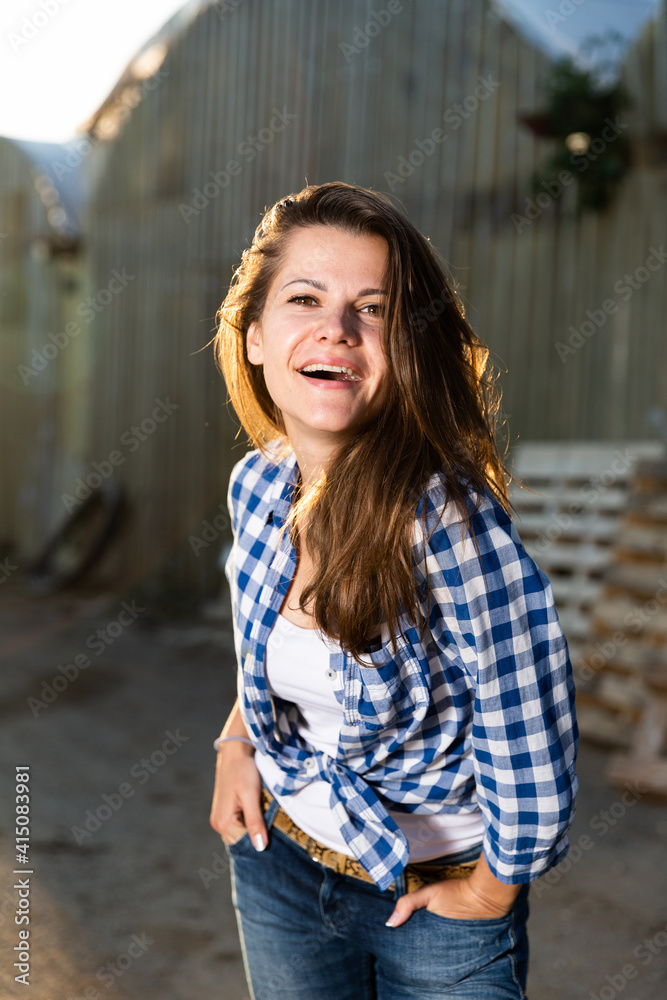 Young woman farmer posing on camera outdoors on background of hothouse buildings..