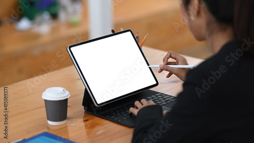 Rear view of businesswoman in suit holding stylus pen writing on digital tablet while sitting at office desk.