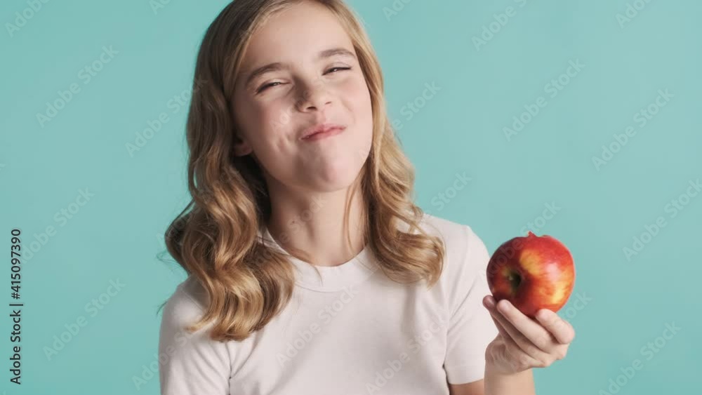 Pretty funny blond teenager girl with wavy hair eating delicious apple and smiling isolated on blue background. Time for snack