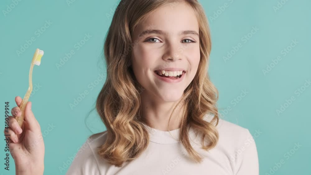 Pretty smiling blond teenage girl with toothbrush checking clean of her teeth on camera over blue background