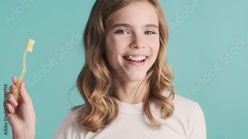 Pretty smiling blond teenage girl with toothbrush checking clean of her teeth on camera over blue background