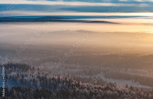Winter landscape with forest, houses, mountains, haze, sky, clouds in the rays of the setting sun