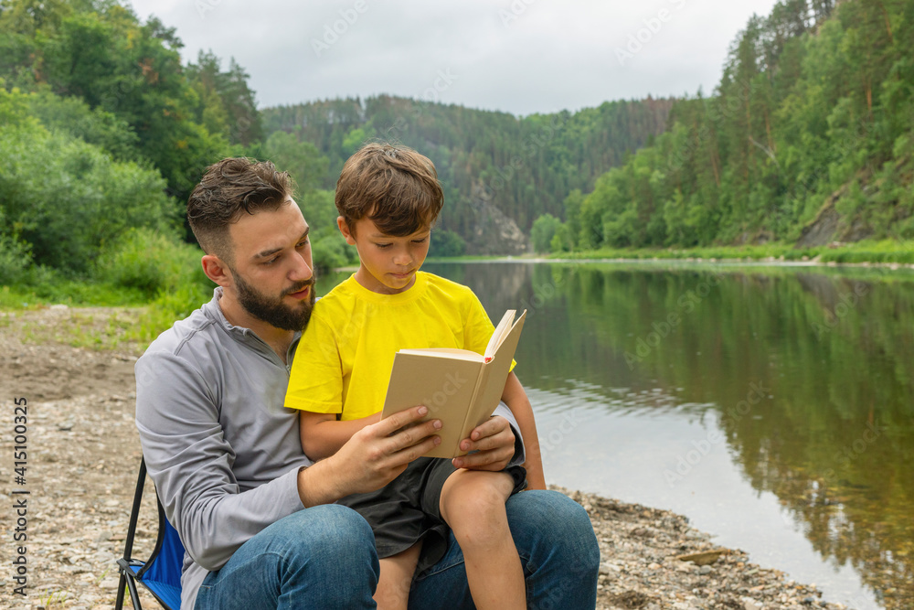 Father and young son reading book together, sitting on beach by river or lake