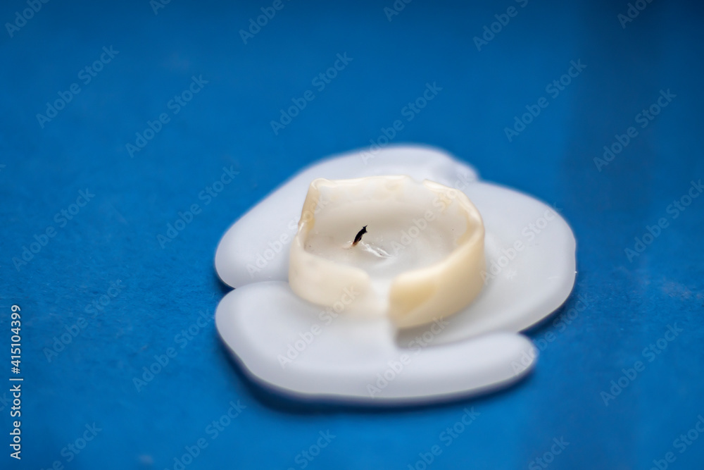 A burnt white, thick candle with molten paraffin wax on blue background. Close-up blurred in soft focus at high magnification. A burnt white, thick candle with molten paraffin wax on blue background. Close-up blurred in soft focus at high magnification.