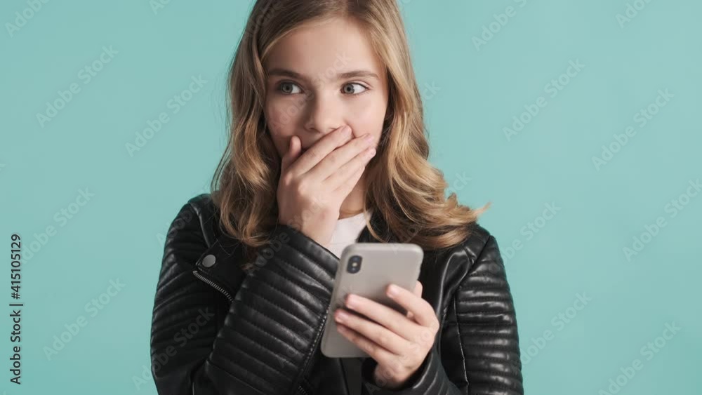 Pretty blond teenager girl getting message from friend on smartphone looking surprised over blue background. Modern technology concept Pretty blond teenager girl getting message from friend on smartphone looking surprised over blue background. Modern technology concept