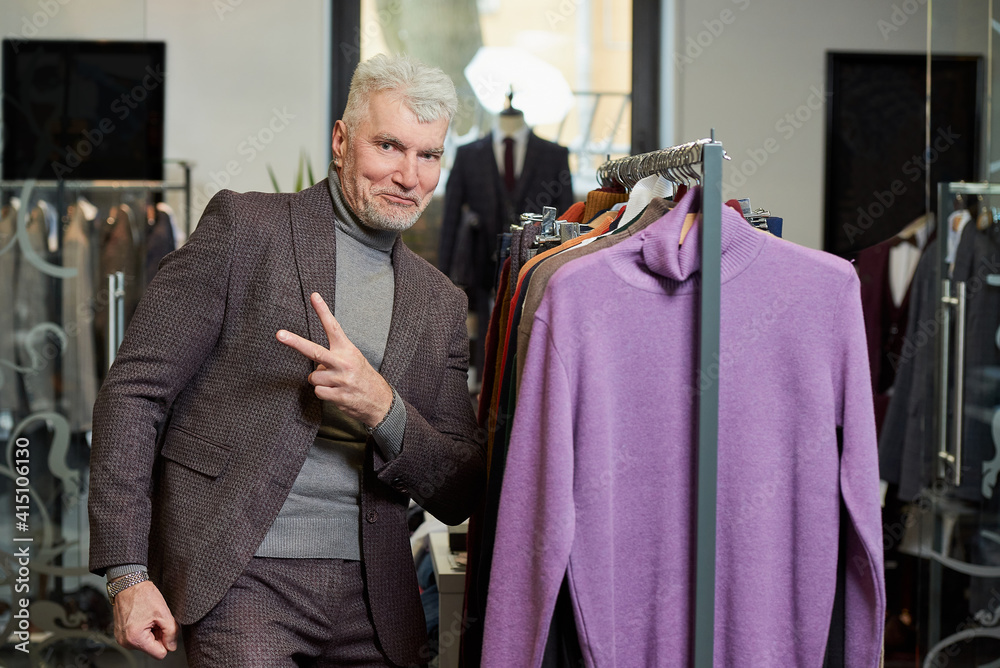 A mature man with gray hair is showing a Victory sign in a clothing store. A happy male customer in a wool suit in a boutique.