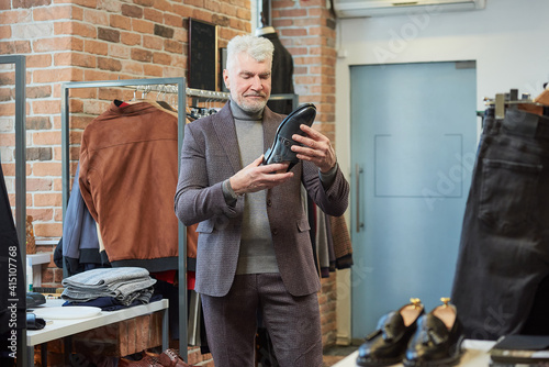 A mature man with gray hair and a sporty physique is choosing shoes in a clothing store. A male customer with a beard wears a wool suit in a boutique