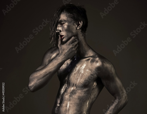 Art guy posing with confident sexy expression, holds finger on lips. Desire concept. Man with streams of sweat on naked chest. Bare torso covered with shimmering silver paint.