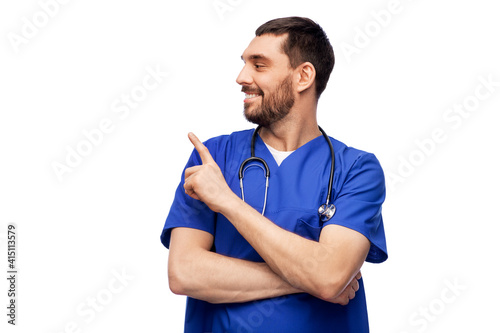 healthcare, profession and medicine concept - happy smiling doctor or male nurse in blue uniform with stethoscope pointing finger to something over white background