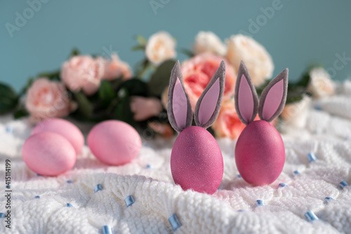 Two pink Easter eggs with bunny ears close-up on a white knitted blanket and a blue background with flowers