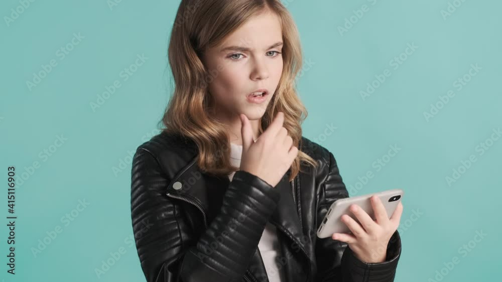 Blond teenager girl dressed in leather jacket watching videos on smartphone looking thoughtful over blue background Blond teenager girl dressed in leather jacket watching videos on smartphone looking thoughtful over blue background