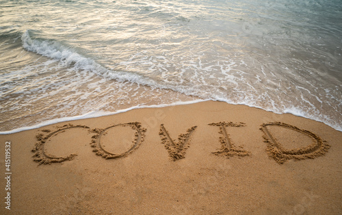 Handwriting COVID on sand and foam wave on beach. Coronavirus concept.