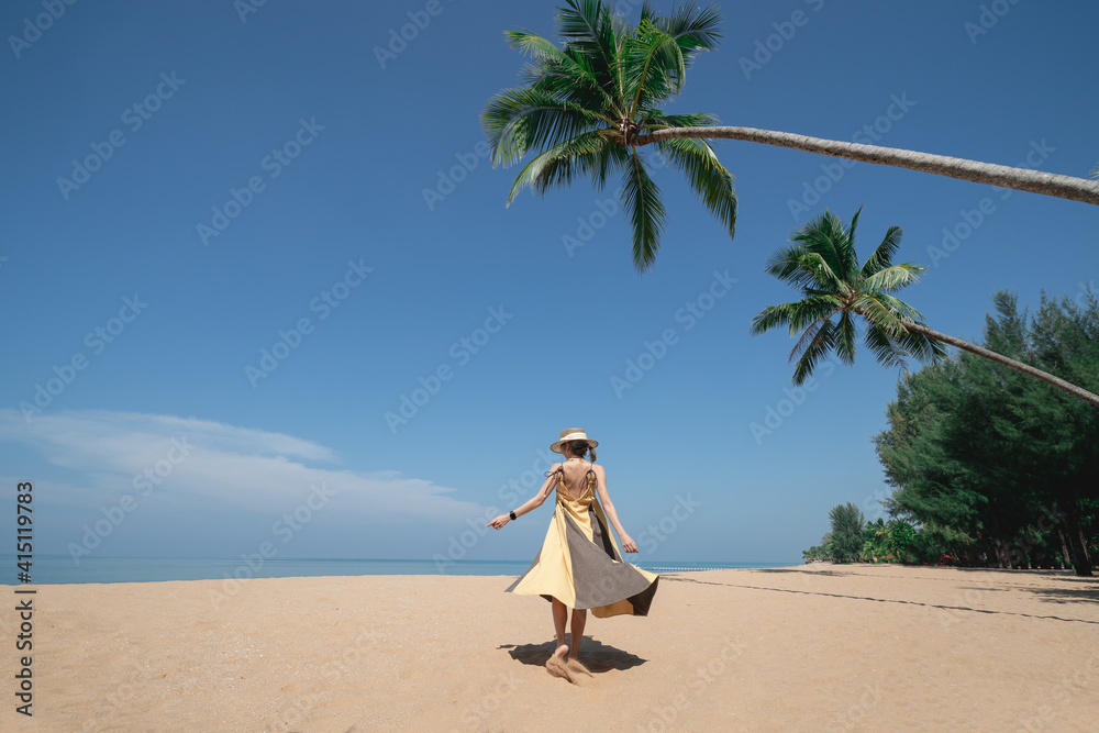 Woman walking under coconut palm tree on the sandy beach with blue sky.