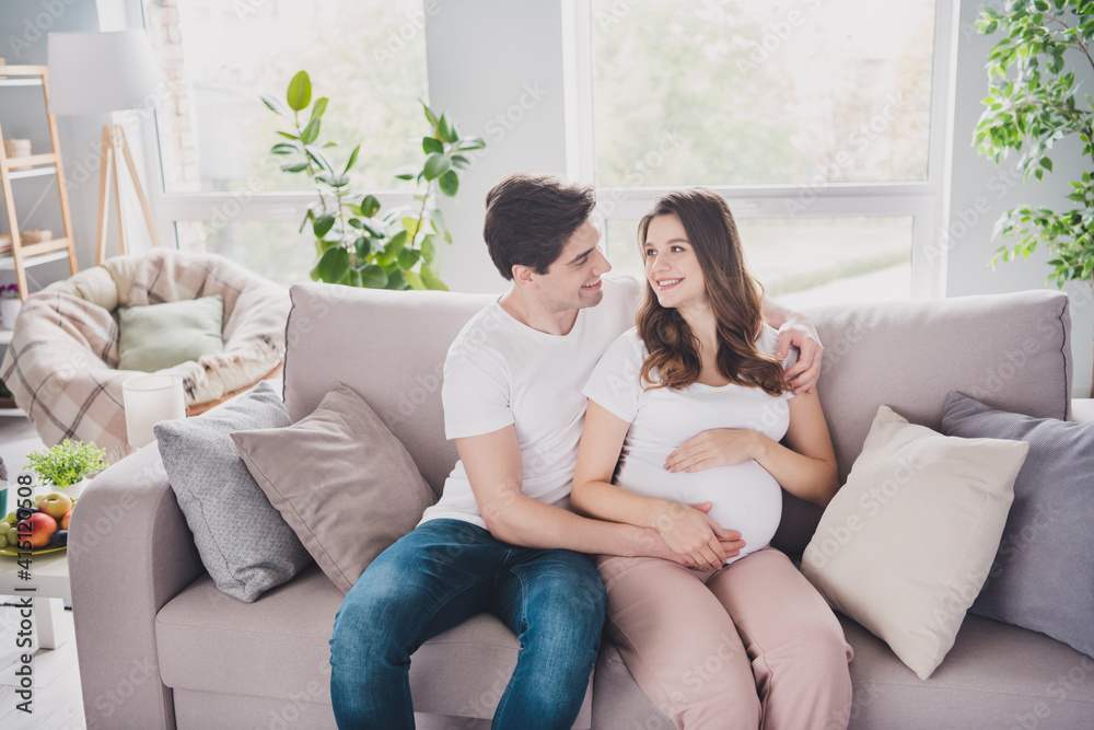 Portrait of adorable affectionate cheerful couple waiting newborn child sitting on divan caressing tummy in light house apartment indoor