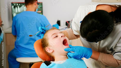 Close up of girl patient lying on stomatological chair with open mouth during dental examination. Stomatologist with mask holding sterilized tools checking teeth health of kid before intervation.