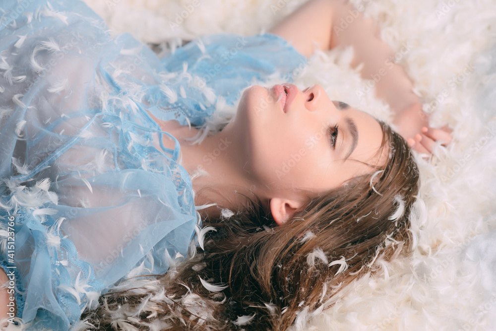 Lady in nightie lay on bed in heap of feathers. Woman in tender pajama relaxing. Girl lay on bed covered with feathers and fluff.