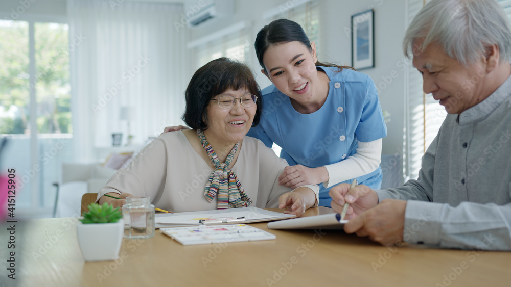 © ChayTee - Attractive young senior asian citizen couple sit at home watercolor painting artwork on desk table with nurse feeling happy in mental health therapy or asia older people quarantine activity lifestyle.