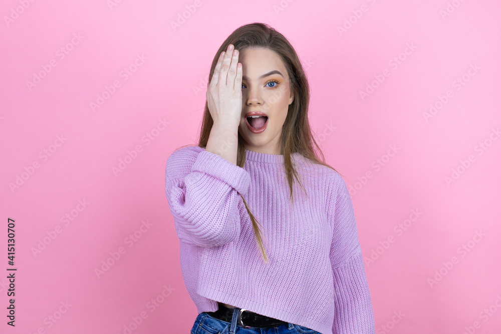 Young pretty woman with long hair standing over isolated pink background covering one eye with hand, confident smile on face and surprise emotion.