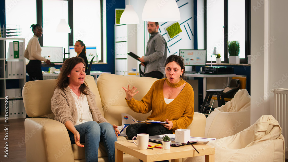 Naklejka premium Stressed busineswomen arguing, yealing at each other during work hours sitting on the couch, while diverse colleagues working scared in background. Women throwing papers, conflicts at work concept