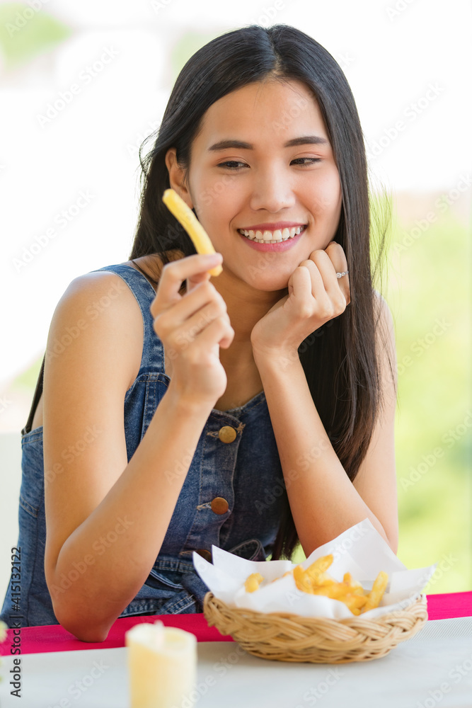 A cute beautiful young Asian woman sitting alone at the fast food shop and holding a piece of french fries and look at it with a happy face A cute beautiful young Asian woman sitting alone at the fast food shop and holding a piece of french fries and look at it with a happy face