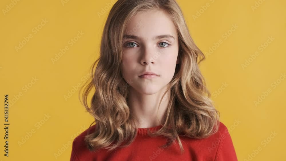 Serious blond teenager girl dressed in red sweater intently looking in camera over yellow background. Confident girl