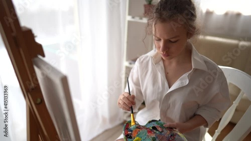 Pretty girl in a white shirt with brush in hand sits near an easel and draws on canvas. Creative teen girl paitning a picture on easel. Girl practising drawing in art school photo