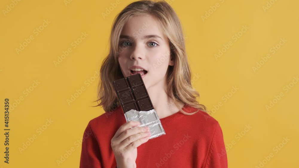 Pretty blond teenager girl dressed in red sweater biting chocolate bar on camera over yellow background