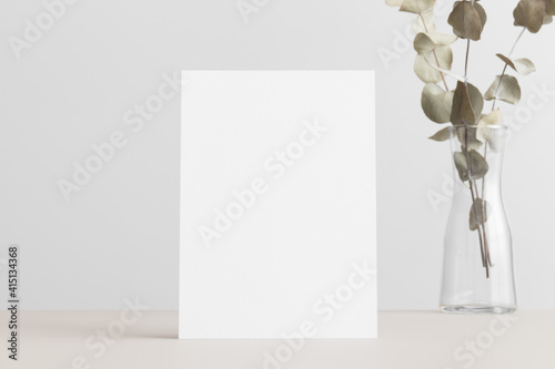 White invitation card mockup with an eucalyptus on the beige table. 5x7 ratio, similar to A6, A5.