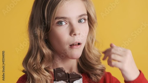 Portrait of attractive blond teenager girl eating chocolate keeping thumb up on camera over yellow background. Tasty expression. Like it gesture