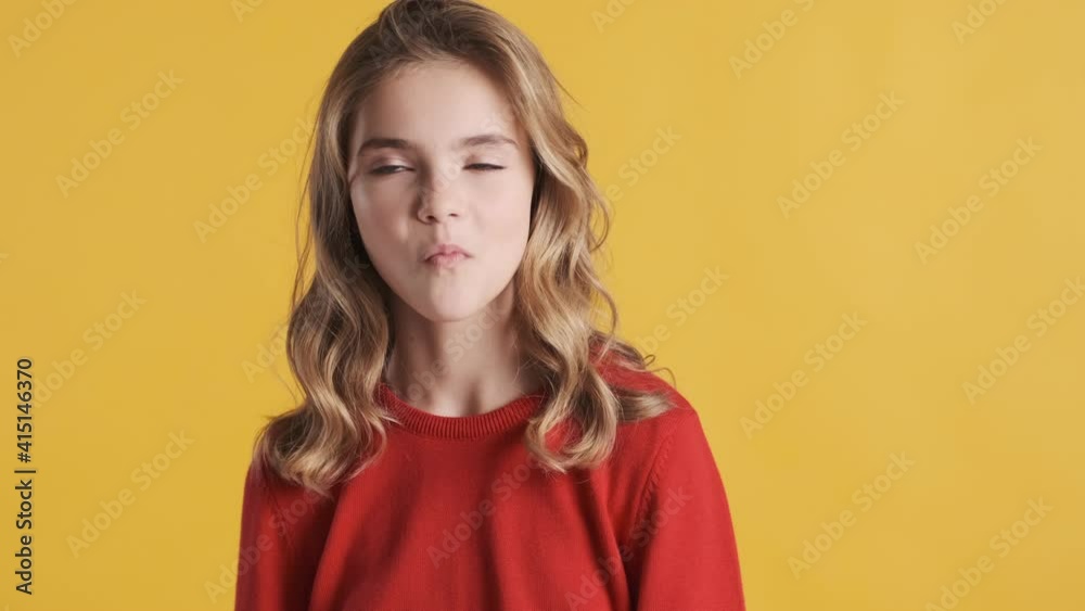 Funny blond teenager girl dressed in red sweater chewing gum on camera over yellow background