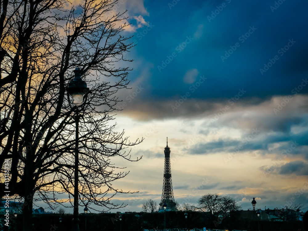 Eiffel Tower at sunrise from a distance with trees and street lamps silhouetted in foreground