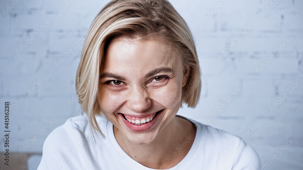 happy blonde woman smiling at camera in bedroom - obrazy, fototapety, plakaty 