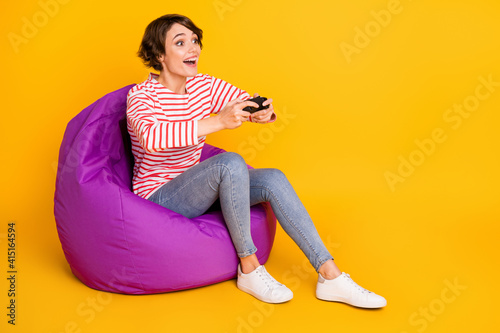 Full size photo of young happy positive good mood girl sit armchair playing playstation isolated on yellow color background