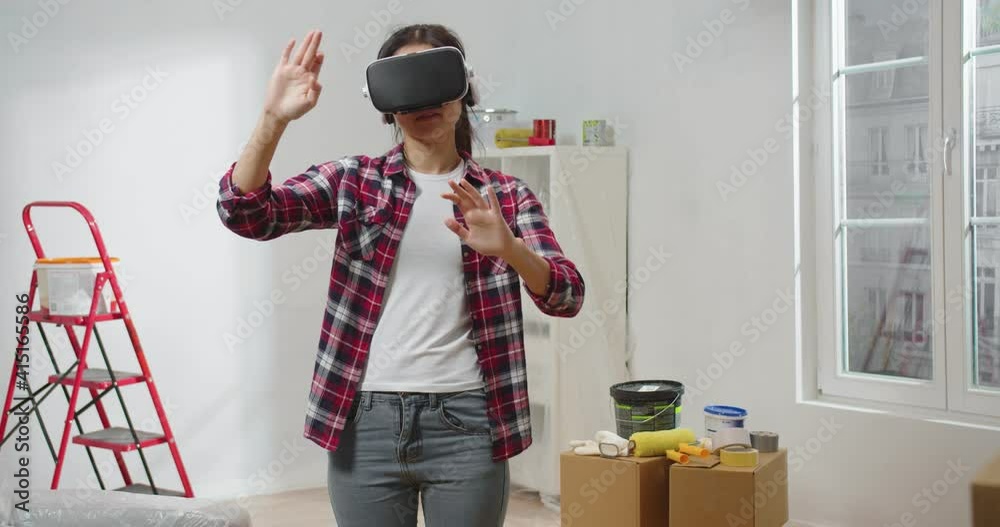 Pretty young woman stand in room during repair works wearing VR glasses using futuristic technology creating room design making moves with hands in air, repair and makeover, virtual reality renovation