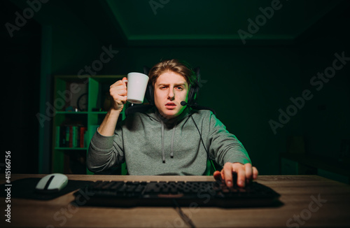 Portrait of a tired gamer in a headset with a cup in his hands playing online games on the computer and looking intently at the camera.