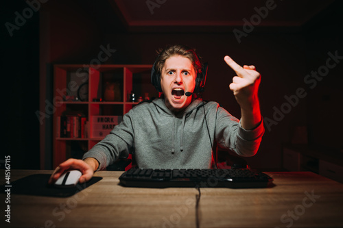 Emotional dissatisfied gamer in a headset angrily looks at camera while playing on the computer and streaming, showing a fuck gesture.Angry guy loses the game and points his middle finger at camera photo