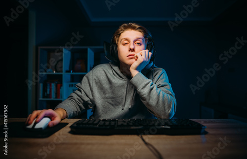 Tired young man in headset playing video games at home on computer in room with blue light and looking at camera with sleepy face.