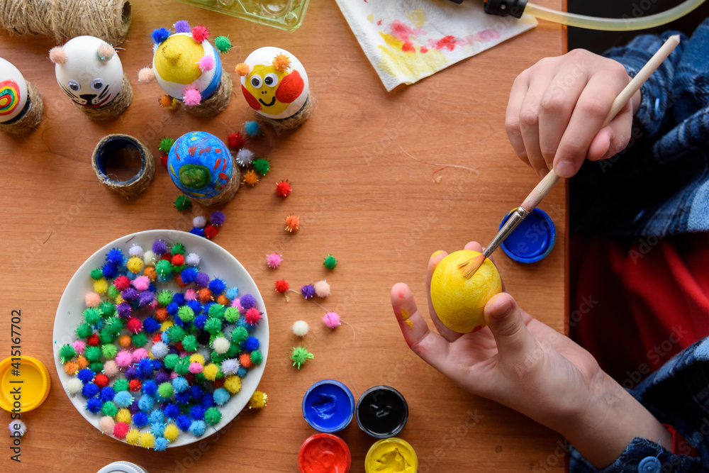 Top view of the desk and the hands of a child who paints festive easter eggs with brushes