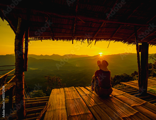 Picture from the back of a woman sitting on wooden porch extending into a high mountain cliff. The sun is setting on the mountain and there is a beautiful warm orange light. The traveling background. © Pang wrp
