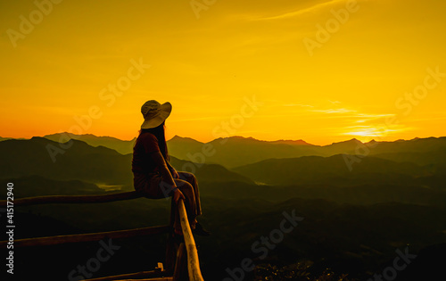 Picture from the back of a woman sitting on wooden porch extending into a high mountain cliff. The sun is setting on the mountain and there is a beautiful warm orange light. The traveling background.