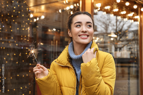 Young woman with burning sparkler near cafe decorated for Christmas