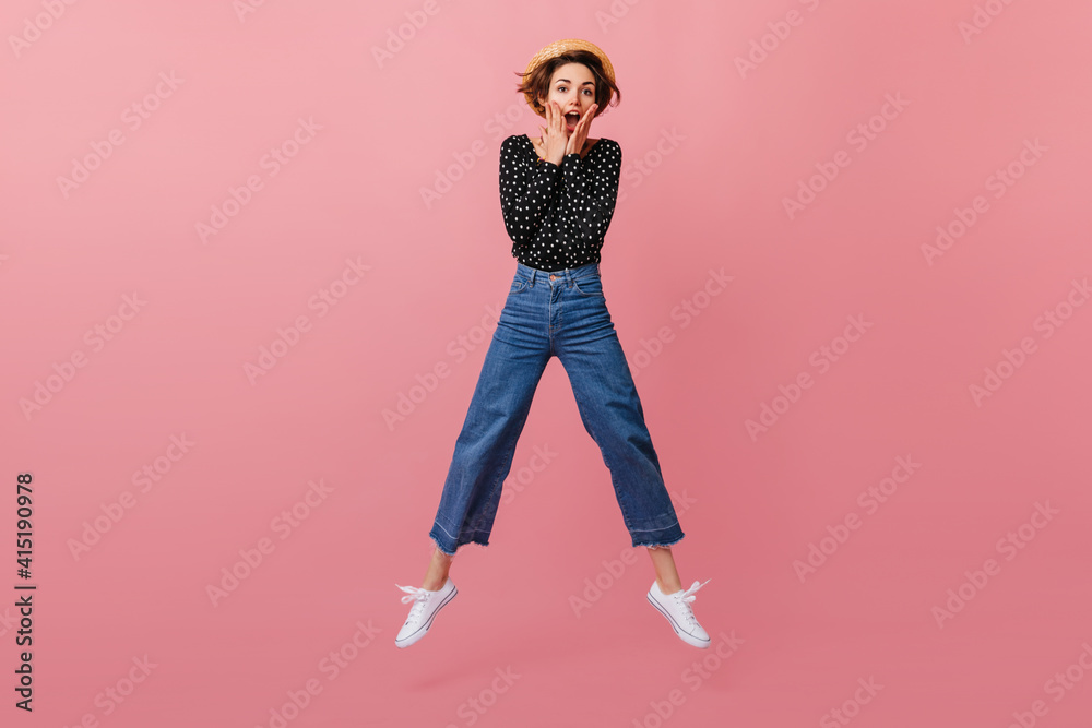 Funny girl in straw hat jumping on pink background. Studio shot of emotional woman in vintage jeans dancing and looking at camera. Funny girl in straw hat jumping on pink background. Studio shot of emotional woman in vintage jeans dancing and looking at camera.
