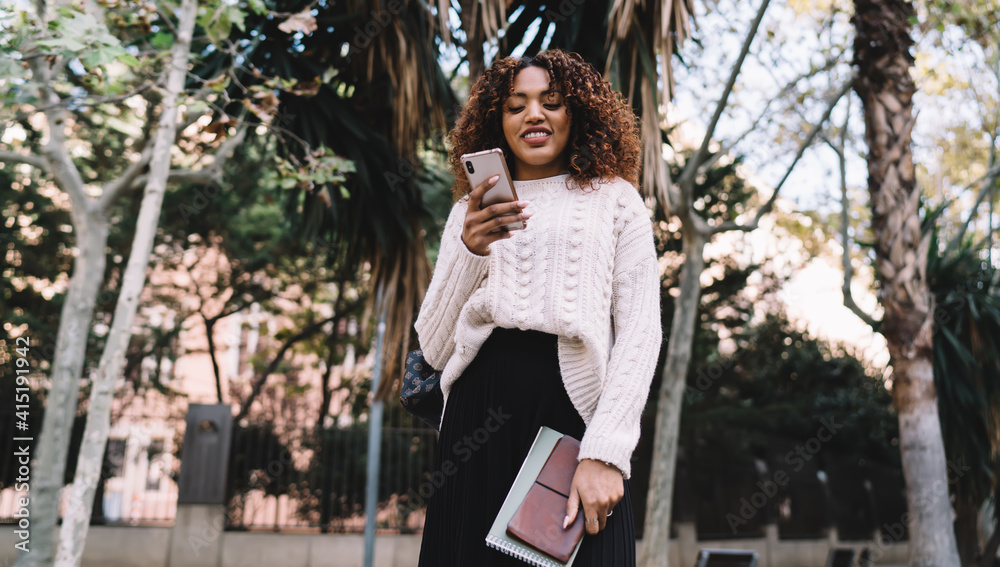 Beautiful dark skinned female in trendy wear reading messages and notification on smartphone spending time in town, positive african american woman blogger satisfied with successful content