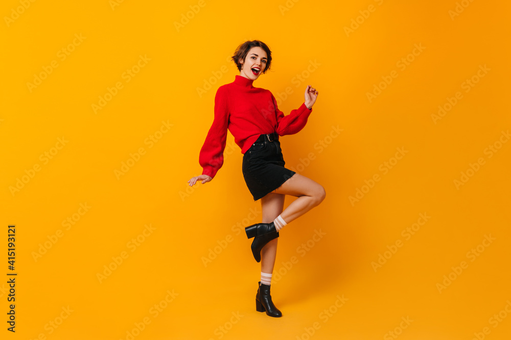 Inspired woman in short skirt dancing on yellow background. Studio shot of blissful brunette girl in red sweater.