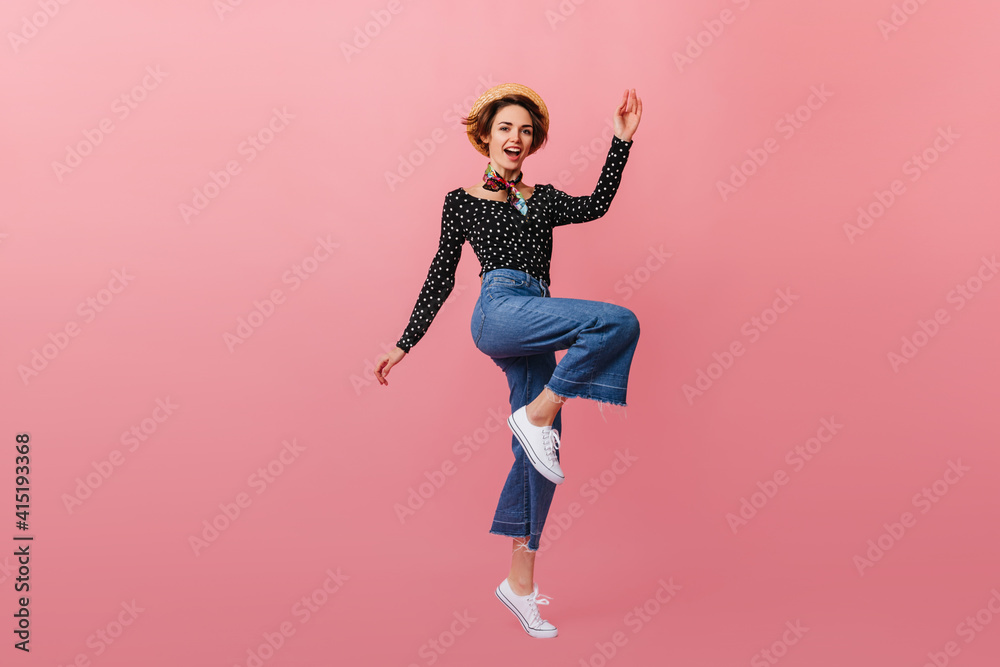 Full length view of jumping woman in straw hat. Studio shot of girl in vintage jeans dancing on pink background.