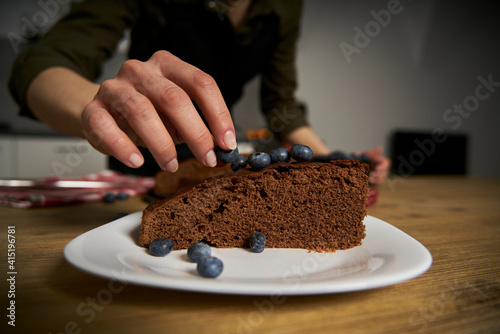 Woman with a passion for cooking decorates a slice of handmade chocolate cake in her home kitchen with blackberries and red currants.