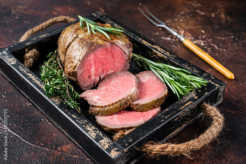 Roast beef round fillet meat in a wooden tray with herbs. Dark background. Top view