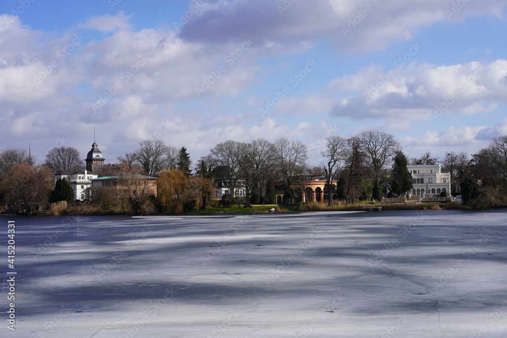 Sonnige winterliche Architekturlandschaft in Potsdam am Heiligen See mit alten Häuser und weißer Eisschicht auf dem Wasser